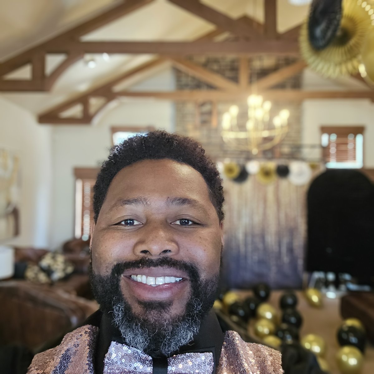 DJ Lil Spoon smiling in a sequin jacket and bow tie in front of a black-and-gold balloon wall.